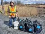 An Adopt-A-Street volunteer wearing an orange vest and standing in front of four garbage bags and a pail.
