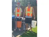 Two men, wearing orange vests, standing. One is holding an orange bucket.
