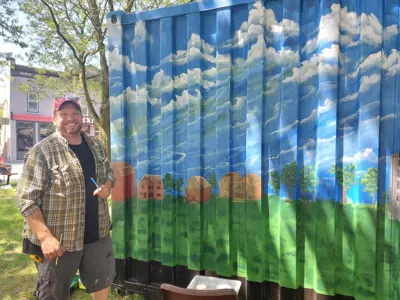 Man smiling and standing in front of a painted mural of sky, trees and grass